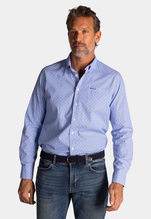 Man met grijze baard, gekleed in een lichtblauw shirt met patroon en blauwe spijkerbroek, voor een effen achtergrond.