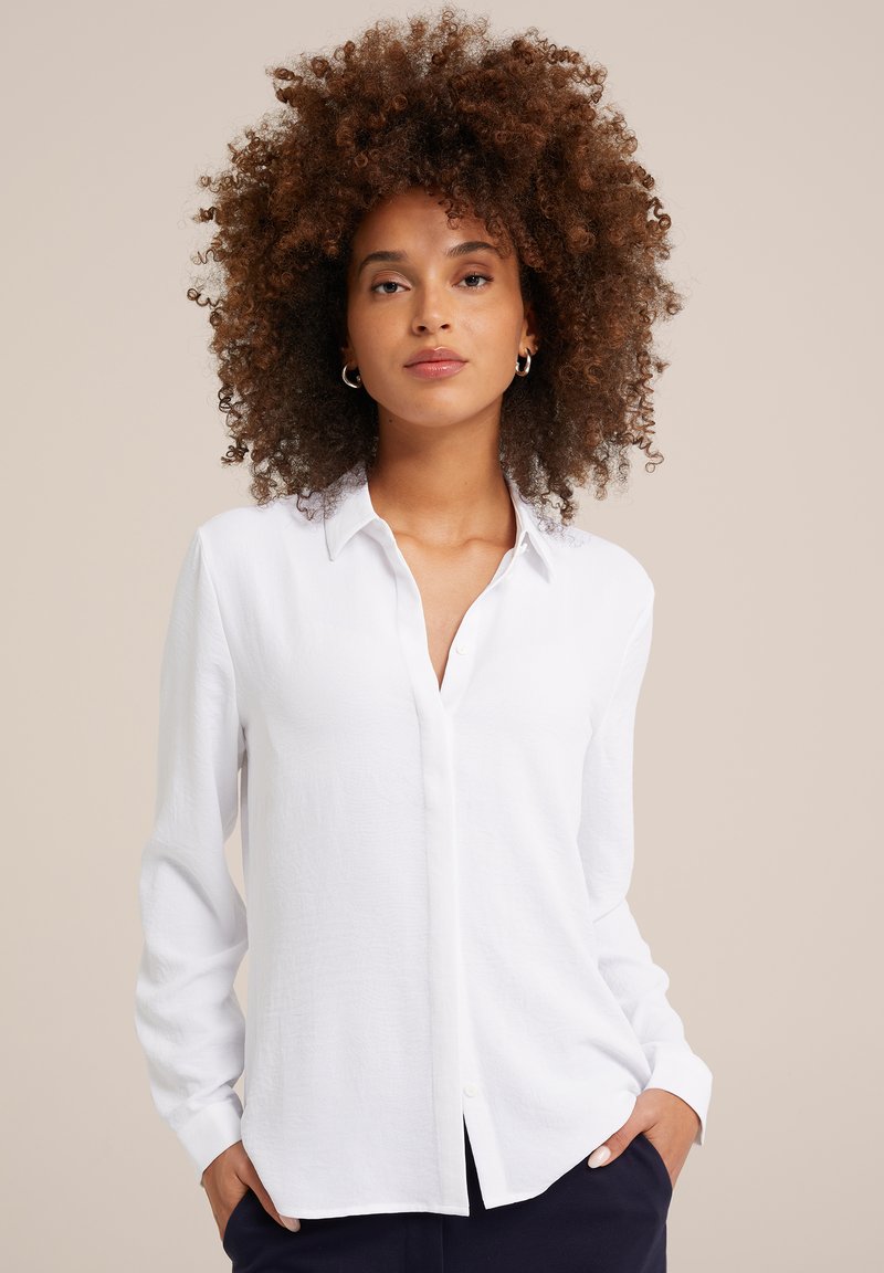 Vrouw met volumineus krullend haar, die zilveren creolen draagt, een witte blouse en een donkere broek aan heeft, staande met haar handen in haar zakken.