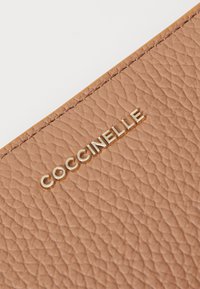 Portafoglio in pelle marrone chiaro con una finitura testurizzata, che presenta un logo "Coccinelle" in oro impresso sulla superficie. I bordi cuciti ne migliorano la durata.