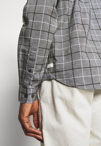 Camicia grigia a quadri con motivo a griglia nero e bianco, polsini con bottoni e un'etichetta sul lato inferiore, indossata con pantaloni chiari.