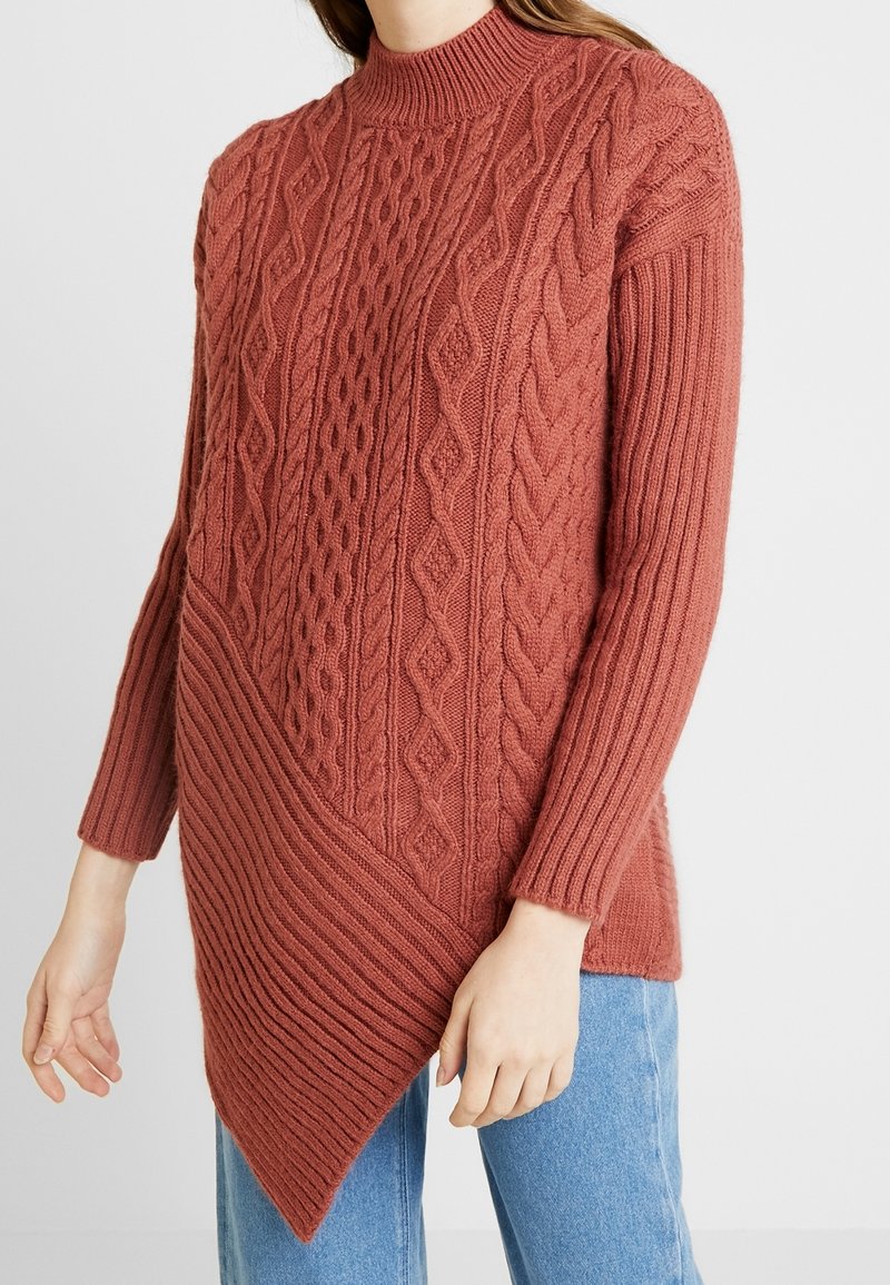Maglione lavorato a maglia color ruggine con collo alto, motivo a treccia, maniche a coste e orlo asimmetrico con texture a coste diagonali.