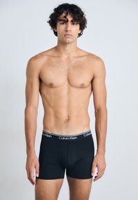 Schwarze Baumwoll-Boxershorts mit einem weichen Bund, der das Logo "Calvin Klein" in Weiß zeigt. Das Modell zeigt eine Frontansicht mit einem glatten, enganliegenden Design.