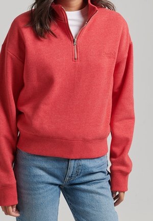 Femme portant un sweat-shirt rouge à quart de fermeture éclair et un jean taille haute bleu clair, debout devant un fond gris uni.