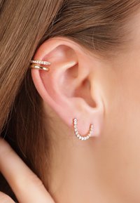 Ohrringe aus Gold mit einem U-förmigen Design, besetzt mit klaren Strasssteinen, und einem zweiten gebogenen Hoop-Ohrring, der durch eine Reihe kleiner Edelsteine akzentuiert wird.