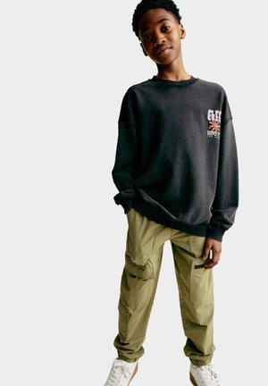 Sweatshirt gris foncé avec un motif sur le devant, coupe décontractée. Pantalons cargo verts olive avec plusieurs poches et poignets élastiques. Des baskets blanches complètent l'ensemble.