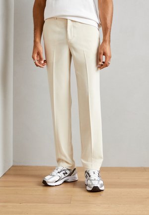 Beige tailored pants with a slim fit, crease detail, and a smooth texture, paired with white athletic shoes featuring gray accents.