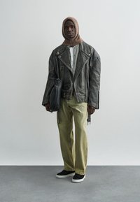 Muškarac stoji, nosi smeđu pletenu kapuljaču, preveliku crnu kožnu jaknu, košulju na kockice, široke zelene hlače i crne cipele, držeći crnu torbu.