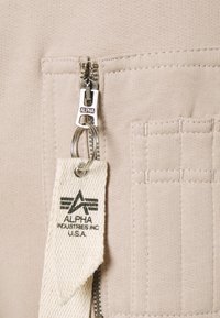 Gros plan sur un tissu beige montrant une fermeture éclair métallique avec une tirette en tissu blanc portant la mention Alpha Industries Inc. U.S.A.