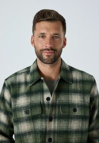 Zaļš un krēmkrāsas krekls ar pārliekamu aizmuguru, divām krūšu kabatām un apkakli, ar strukturētu audumu un tumšiem pogām.