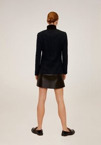 Schwarzer strukturierten Blazer mit Kragen, kombiniert mit einem schwarzen Leder-Minikleid. Modell trägt schwarze Loafer, von hinten betrachtet.