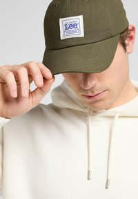 Gorra de béisbol verde oliva de algodón, con un parche blanco cuadrado que tiene texto azul. El portador lleva puesta una sudadera neutra visible.
