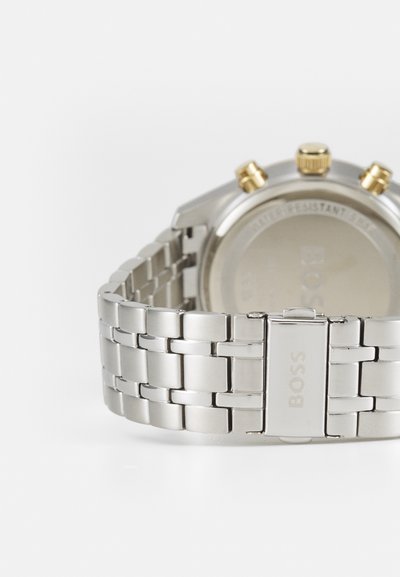 Montre en acier inoxydable argenté avec un bracelet texturé, un boîtier rond et des accents dorés sur les poussoirs du chronographe. Fond de boîtier transparent avec détail du logo.