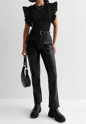 Woman dressed in black ruffled sleeveless top, high-waisted leather pants, black belt, chunky boots, and holding a black handbag.