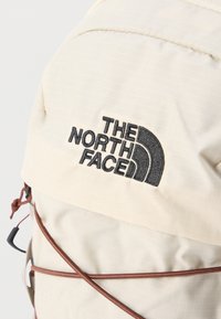 Σακίδιο πλάτης σε κρεμ απόχρωση με υφή, που διαθέτει μαύρο λογότυπο στο μπροστινό μέρος και καφέ ελαστικά κορδόνια για στερέωση αντικειμένων.