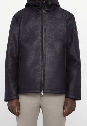 Veste noire texturée à capuche avec fermeture éclair frontale et poches latérales portée sur un pantalon beige clair, montrée de la taille au cou.