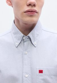 Ανοιχτό μπλε πουκάμισο με κουμπιά, κατασκευασμένο απ ό βαμβάκι, με γιακά, λευκά κουμπιά και τσέπη στο στήθος που φέρει μια μικρή κόκκινη ετικέτα "HUGO".