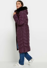 Lang lila dunfrakke med hætte og kunstig pelskant, med quiltet tekstur, sidelommer med lynlås og trykknapper. Båret over blå jeans og hvide sneakers.