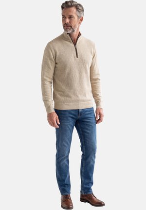Uomo di mezza età che indossa un maglione beige con zip a quarti, jeans blu e scarpe di pelle marrone, in piedi e che guarda leggermente di lato.