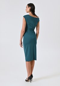 Teal-farbenes, tailliertes Kleid mit kurzen Ärmeln, einem Rückenzipper und einer knielangen Silhouette, kombiniert mit schwarzen High-Heels. Texturiert.