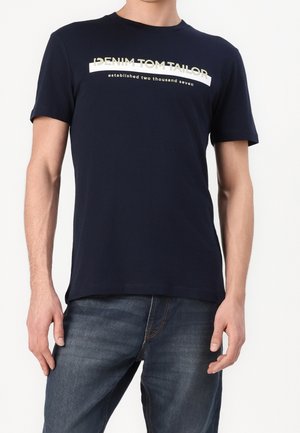 Homme portant un t-shirt en denim bleu marine Tom Tailor et un jean bleu, debout devant un fond blanc uni.