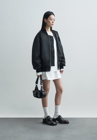 Czarna kurtka bomberka na białej sukience, w zestawie z białymi skarpetkami do kostek i brązowymi butami. Całość dopełnia mała czarna torebka z ćwiekami.