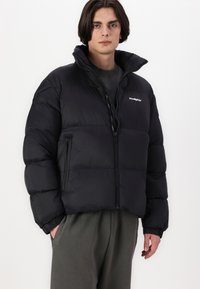 Svart pufferjakke med høy krage, glidelås foran og sidelommer. Den har et quilted design med matt finish og elastiske mansjetter.
