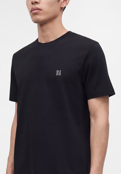 Homme portant un t-shirt noir uni à manches courtes avec un petit logo gris D2 sur la poitrine gauche sur fond blanc.
