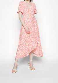 Vestido envolvente floral rosa con mangas cortas abullonadas, escote en V y dobladillo asimétrico. Presenta detalles en dorado y una textura de tela ligera y fluida.