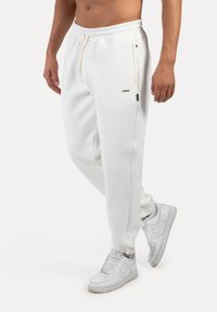 Bílé tepláky s stahovací šňůrkou v pase, zúženými manžetami a bočními kapsami. V kombinaci s bílými teniskami. Hladká bavlněná tkanina.