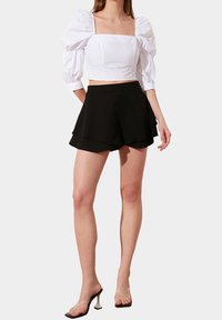 Bílý crop top s nafouklými rukávy, kombinovaný s černými vrstvenými šortkami a transparentními sandály na podpatku. Hladká tkanina a strukturované designové prvky.