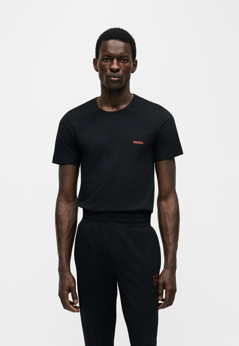 Mann trägt ein schwarzes kurzärmeliges T-Shirt mit rotem „HUGO“-Logo und dazu passende schwarze Hose vor einem einfarbigen hellen Hintergrund.