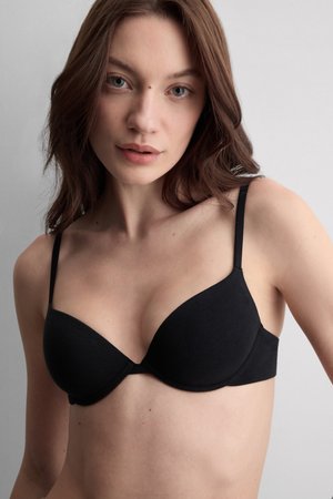 Frau mit heller Haut und braunen Haaren, die einen schwarzen BH trägt, blickt nach vorne vor einem einfarbigen grauen Hintergrund.