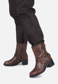 Botas de tobillo de cuero marrón con un tacón grueso, que presentan cremalleras laterales, correas decorativas y acentos de metal. Diseño clásico de punta redonda.
