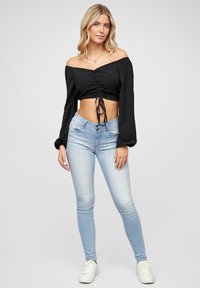 Fekete crop top, off-shoulder dizájnnal, ráncolt elülső résszel és megkötős részlettel. Világoskék karcsú farmerral és fehér sportcipővel párosítva.