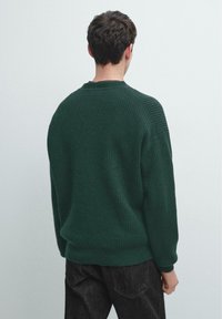 Dunkelgrüner gestrickter Pullover mit geripptem Muster, Rundhalsausschnitt und überschnittener Schulterpartie. Sichtbare Nähte und lässige Passform, kombiniert mit dunklen Jeans.