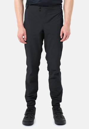 Mann, der schwarze, eng anliegende Outdoor-Hosen und schwarze Sportschuhe trägt, steht vor einem einfarbigen weißen Hintergrund.