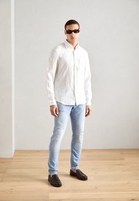 Weißes Leinenhemd mit langen Ärmeln, hellblaue Jeans mit Rissen, schwarze Slipper und dunkle Sonnenbrille. Steht auf einem Holzboden vor einer hellen Wand.