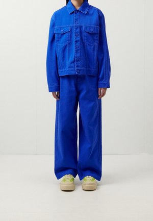Person iført en koboltblå jakke med knapper og matchende vidde bukser kombineret med beige og grønne sneakers, står foran en ensfarvet baggrund.