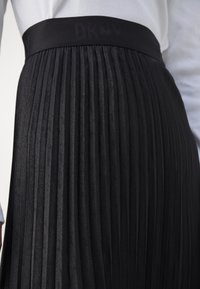 Gros plan d'une jupe plissée noire avec une large ceinture arborant un logo DKNY discret, portée avec un haut blanc.
