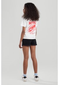 Weißes T-Shirt mit rotem Grafikdruck, der den Text "Montauk" und ein Schuhdesign auf der Rückseite zeigt, kombiniert mit schwarzen Shorts und weißen Sneakers.