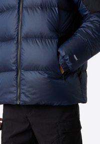 Mörkblå pufferjacka med horisontell sömnad, dragkedjestängning, elastiska ärmmuddar och en '700'-detalj på ärmen. Buren över svarta byxor.