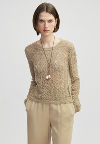 Beige Strickpullover mit einem strukturierten Zickzackmuster, runden Ausschnitt und langen Ärmeln, kombiniert mit hellbeigen Hosen.