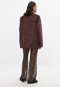 Blazer oversize in suede marrone con una finitura texture, abbinato a pantaloni in pizzo nero trasparente con motivi floreali.