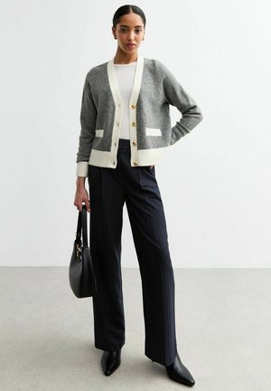 Grijze cardigan met crème accenten en zakken, voorzien van gouden knopen. Gecombineerd met marineblauwe streepjesslacks en zwarte puntige laarzen.