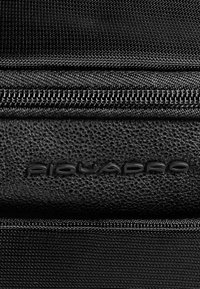 Zwarte gestructureerde tas met een glad leren accent met het verdiept aangebrachte "Piquadro"-logo en een zichtbare metalen rits langs de bovenrand.