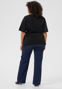 Camisa negra de manga corta con detalle de lazo lateral, combinada con jeans rectos de mezclilla azul oscuro, que presentan una textura de mezclilla clásica y bolsillos traseros.