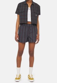 Geschnittenes karriertes Hemd in Marineblau, mit kurzen Ärmeln und zwei Vorderaschen; kombiniert mit karrierten Shorts, elastischem Bund und gelben Turnschuhen.