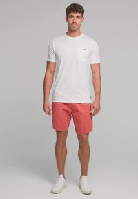 Bărbat în picioare purtând un tricou alb cu mânecă scurtă și buzunar, pantaloni scurți corali și adidași albi, pe un fundal gri simplu.