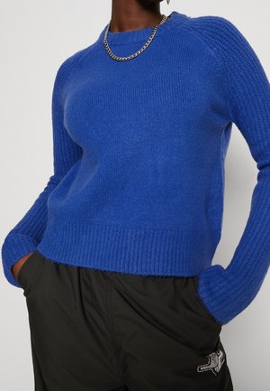Personne portant un pull en maille bleu vif avec des manches texturées et un pantalon noir, les mains dans les poches, accessoirisée d’un collier chaîne argenté.