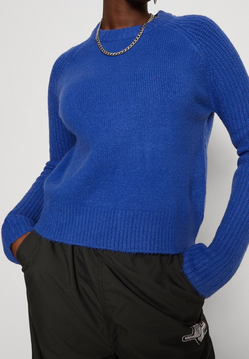 Personne portant un pull en maille bleu vif avec des manches texturées et un pantalon noir, les mains dans les poches, accessoirisée d’un collier chaîne argenté.